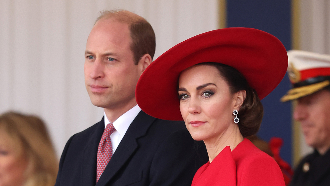 Prince WIlliam, a white man in a tuxedo and Kate Middleton, a white woman with brown hair wearing a red dress and hat, staring in the distance.
