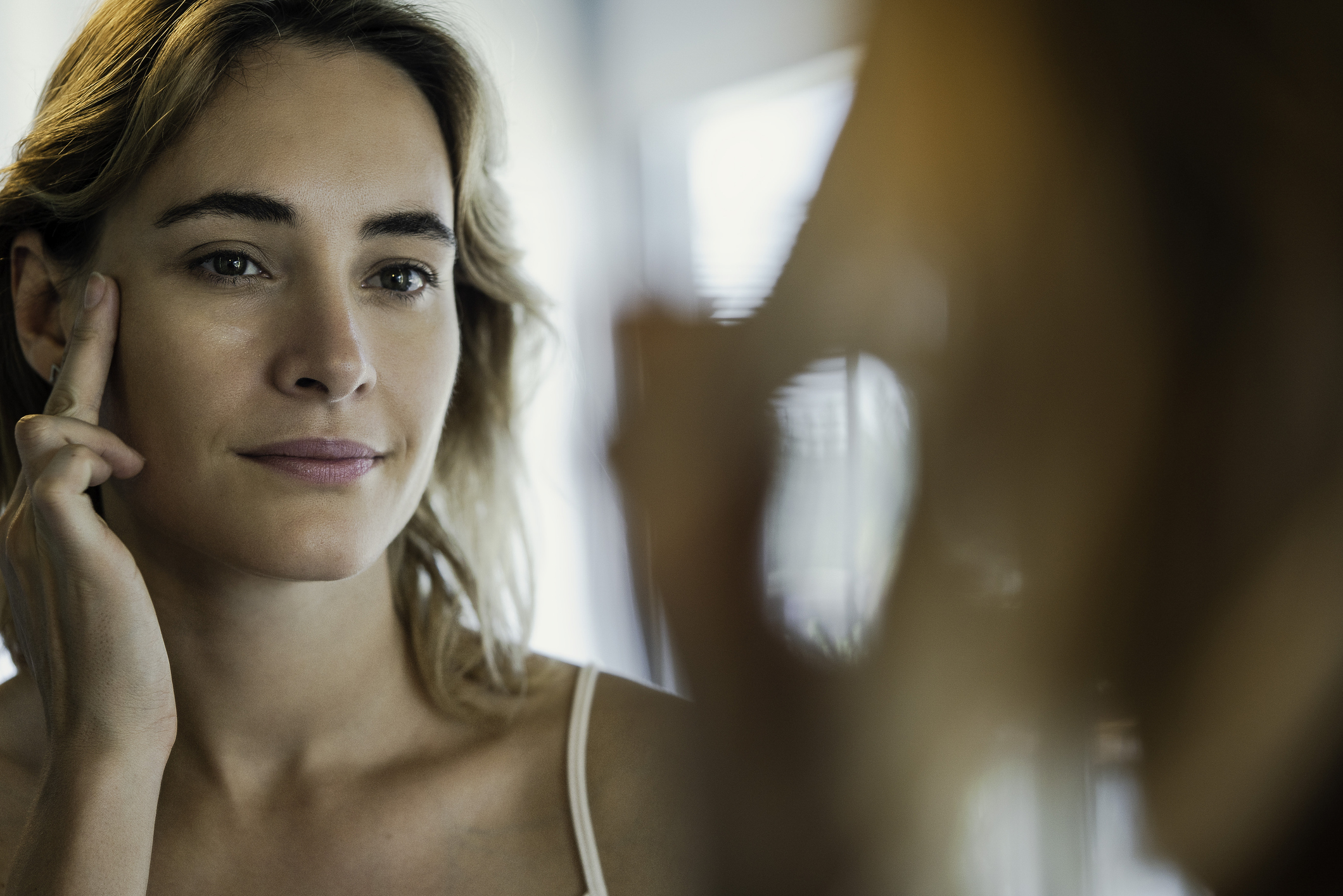 A woman looking at herself and touching her face in the mirror.