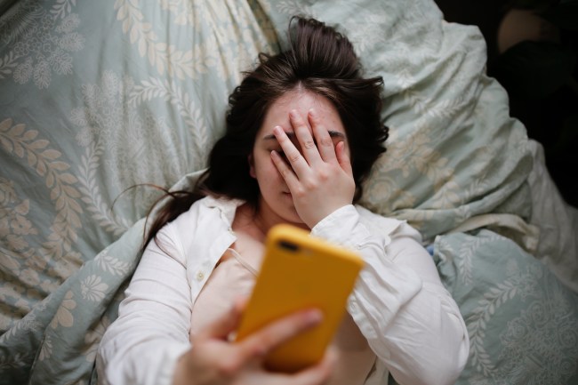 Young woman in home bedroom on bed with phone. Woman covering her eyes with her hand