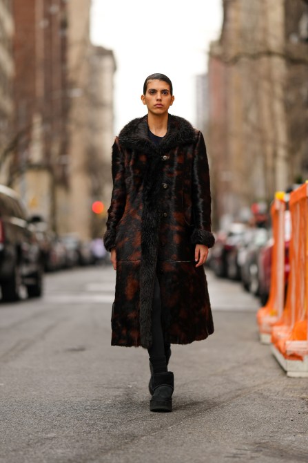 A guest wears a black pullover, a long fur coat, black tights, and black platform Ugg boots during NYFW on February 12, 2023 in New York City.